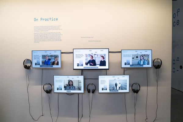 Image of five screens with headphones on gallery wall, displaying digital interviews from the exhibition.