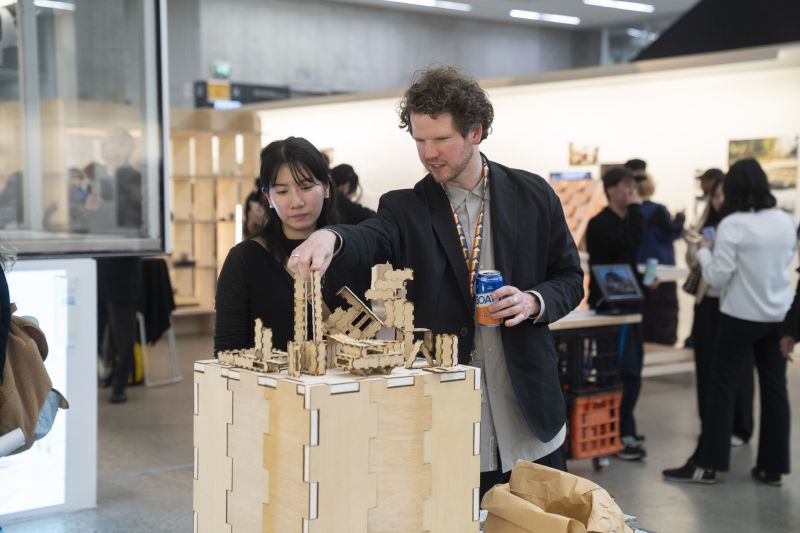 Two people stand behind a series of models inside a busy white-wall gallery space. 