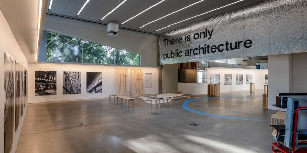 Concrete exhibition gallery dimly lit with a metallic silver banner proclaiming 'there is only public architecture'
