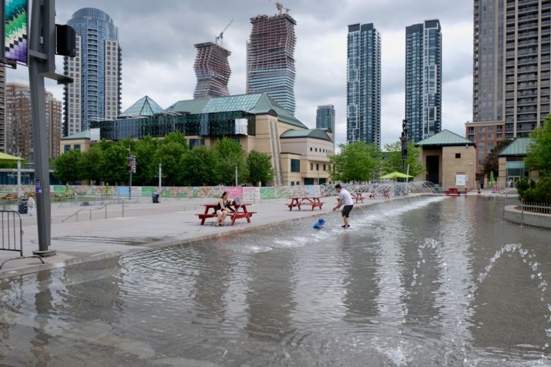 A photograph of a child and parent playing in a water fountain, in front of a cityscape.