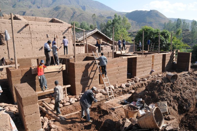 workers construct rammed earth walls on site. In the foreground are piles of loose earth and rocks, and in the background are mountains.