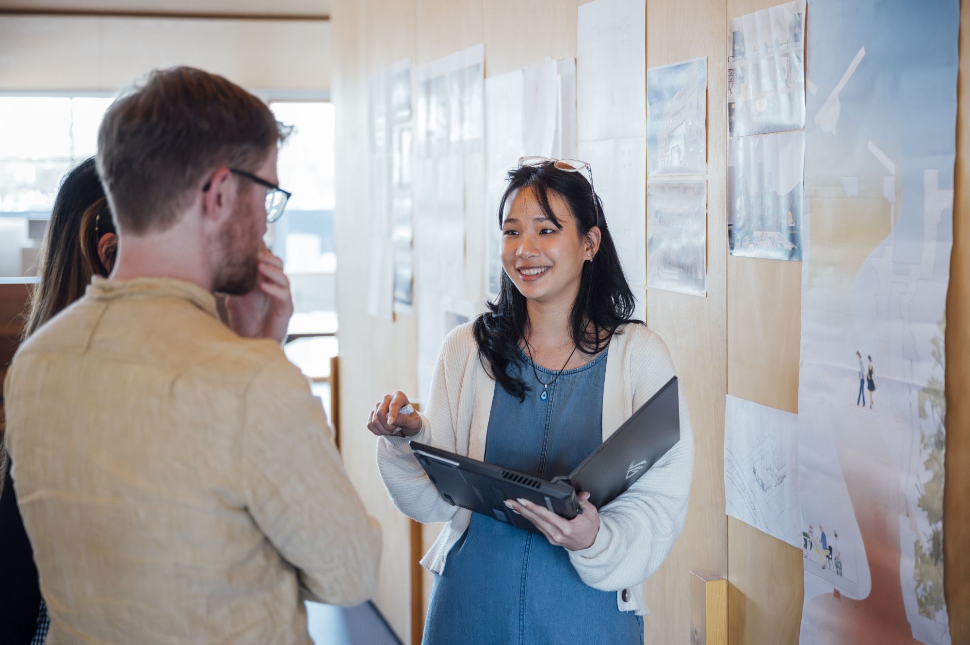 A person with long hair holds a laptop and smiles as she talks to other people in front of a poster